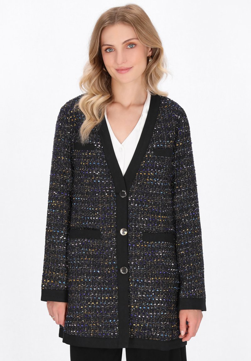 Zwarte tweed cardigan met veelkleurige spikkels, v-hals, knoopsluiting, twee zakken en zwarte biezen. De lengte reikt tot net onder de heupen.