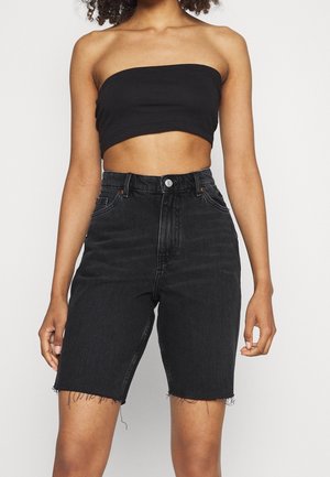 Frau trägt ein schwarzes trägerloses Crop-Top und schwarze High-Waist-Denim-Shorts mit ausgefransten Säumen, steht vor einem einfarbigen Hintergrund.