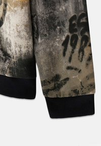 Giacca di cotone in tonalità marrone, nero e grigio. Presenta un polsino nero a costine e una superficie testurizzata con dettagli di impronte di mani e motivi.