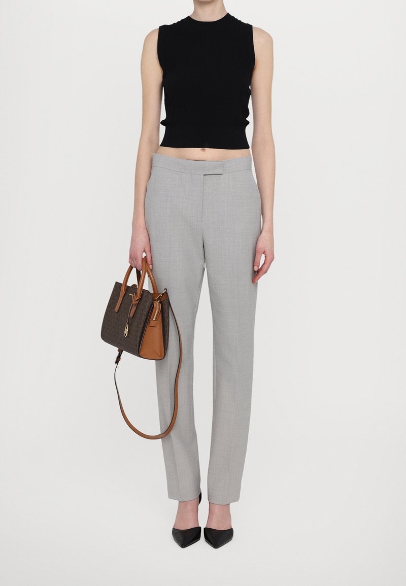 Personne portant un crop top noir sans manches, un pantalon gris clair sur mesure, des chaussures noires pointues et tenant un sac à main brun à motif sombre.