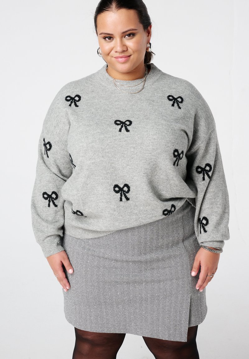 Femme souriante portant un pull gris avec des motifs de nœuds noirs, une jupe grise à motifs et des collants noirs, debout devant un fond uni.