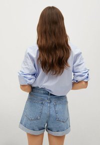Mujer con largo cabello castaño, vistiendo una camisa de rayas azul clara con mangas remangadas y pantalones cortos de mezclilla de tiro alto, de espaldas a la cámara.