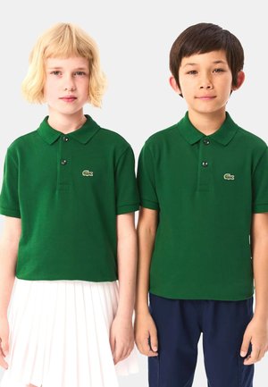 Polo verde con un tessuto texturizzato, dotato di colletto e tre bottoni, indossato da due bambini. Uno indossa una gonna plissettata bianca, l'altro pantaloni scuri.