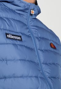 Blå vadderad jacka med hög krage, med svart "ellesse"-logotyp och mångfärgat emblem. Mjuk textur och quiltad design.