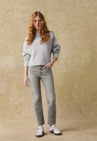 Hellgrauer gestrickter Pullover mit lockeren Ärmeln und rundem Halsausschnitt, kombiniert mit hellgrauen Jeans und weißen Sneakern mit braunen Sohlen.