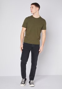 Olīvu zaļš īsām piedurknēm T-krekls ar apaļu kakla izgriezumu, apvienots ar melnām šaurām džinsām un melniem sporta apaviem ar baltām zoles. Vienkāršs dizains, bez rakstiem.