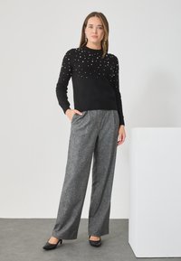 Czarny sweter dzianinowy z perłowymi detalami, w zestawieniu z szarymi spodniami o szerokich nogawkach. Modelka ma na sobie czarne szpilki w szpic. Proste tło.