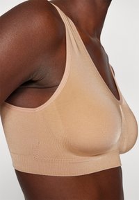 Beige saumaton bralette, jossa on syvä V-pääntie, leveät olkaimet ja sileä, venyvä rakenne. Mallissa on muotoiltu siluetti.