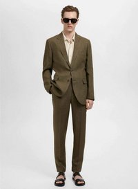 Bruin op maat gemaakt pak van gestructureerde stof, gecombineerd met een crèmekleurig shirt en zwarte zonnebril. Model draagt zwarte sandalen.