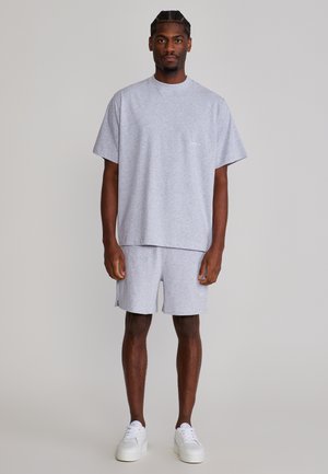 Uomo in piedi che indossa una maglietta grigia oversize e pantaloncini abbinati con sneakers bianche, su uno sfondo chiaro e semplice.