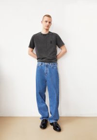 Grijze katoenen t-shirt met een zwart lintlogo, gecombineerd met blauwe losse denim jeans en zwarte leren schoenen. Model staat tegen een witte muur.