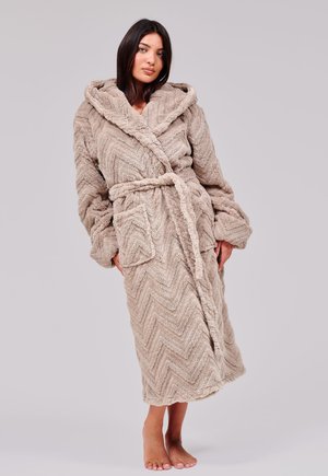 Vrouw staat blootsvoets en draagt een lange, gestructureerde beige badjas met kap en riem die in de taille is geknoopt tegen een effen achtergrond.