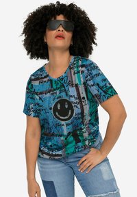 T-shirt à manches courtes de couleur bleue avec des motifs abstraits noirs et verts, présentant un graphique central de smiley et une coupe ample.