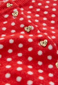Punainen kangas, jossa on valkoisia pilkkuja ja kultaisia sydämenmuotoisia koristeita. Kuvio näyttää pehmeältä ja pörröiseltä, mikä korostaa leikkisää muotoilua.