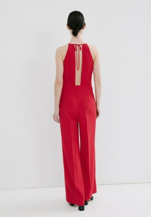 Femme aux cheveux foncés attachés en chignon, portant une combinaison rouge sans manches avec un dos ouvert et haut noué au cou, debout contre un mur blanc uni.