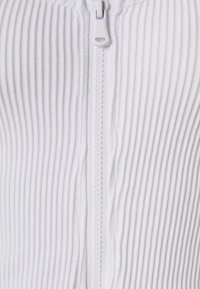 Indumento bianco con zip, caratterizzato da una texture a coste, con una zip semplice e materiale morbido. Inquadratura ravvicinata che mette in evidenza il tessuto dettagliato e la zip.