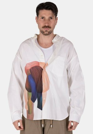 Uomo con capelli corti e scuri e baffi indossa una camicia bianca larga con disegno astratto colorato sopra una canottiera bianca e pantaloni beige.
