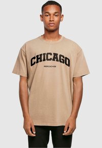 Beige puuvillat-paita, jossa on rohkeat mustat kirjaimet "CHICAGO" ja "MERCHC•DE", lyhyet hihat ja rento leikkaus.