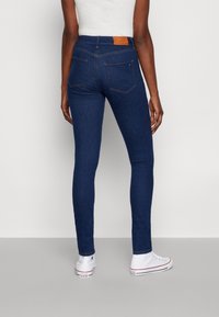 Jeans oscuros de corte slim con bolsillos traseros, llevados por una persona de pie, combinados con zapatillas Converse blancas de caña alta.