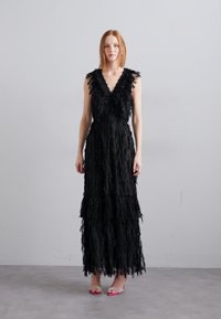 Robe noire longue avec des détails en franges superposées, décolleté en V profond et sans manches, complétée par des sandales à brides roses.