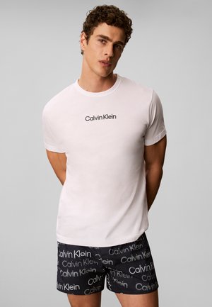 Άνδρας που φοράει λευκό μπλουζάκι Calvin Klein και μαύρα σορτς με επαναλαμβανόμενο λογότυπο Calvin Klein, στέκεται με τα χέρια πίσω από την πλάτη μπροστά σε γκρι φόντο.