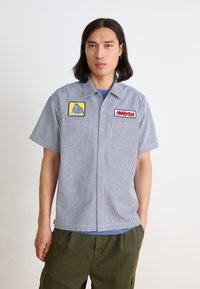 Kortermet stripet skjorte i blått og hvitt, med to broderte merker, ett gult med fjelldesign og ett rødt.