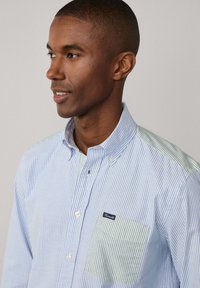 Camicia a righe con bottoni, caratterizzata da righe verticali blu e bianche, tessuto verde a contrasto sulla spalla e una tasca sul lato sinistro del petto.