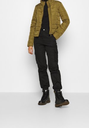 Osoba ubrana w oliwkowo-zieloną pikowaną kurtkę, czarny golf, czarne spodnie typu cargo i czarne sznurowane botki stoi na tle gładkiej białej ściany.