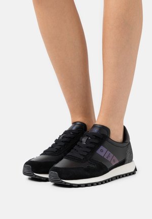Jambes portant des baskets noires à lacets avec des bandes latérales violettes, du daim noir, des détails en cuir, et des semelles blanches sur un fond blanc uni.