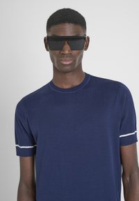 Maglietta a maglia blu navy con maniche corte e accenti a righe bianche sui polsini, abbinata a occhiali da sole neri angolari.