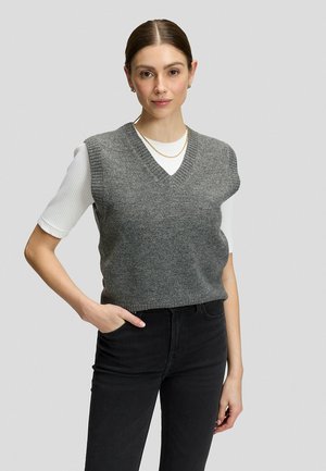 Donna con capelli castani divisi al centro indossa un gilet grigio senza maniche sopra una maglia bianca e jeans neri, in piedi contro uno sfondo semplice.