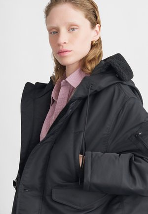 Svart isolert jakke med en stor hette, glidelås og flere lommer; båret over en lys rosa teksturert skjorte.