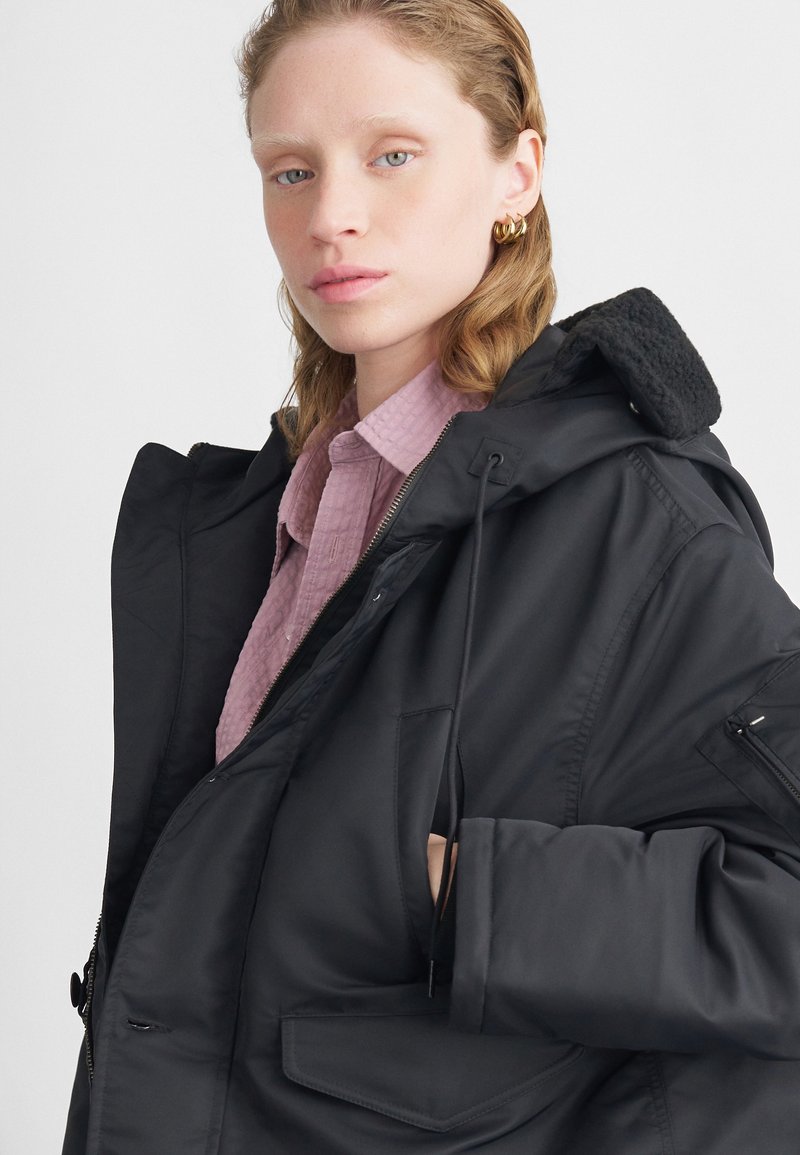 Melna izolēta jaka ar mīkstu kapuci, rāvējslēdzēja aizdari un vairākiem kabatām; valkāta virs gaiši rozā teksturētas krekla.