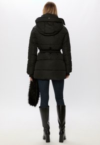 Sort pufferjakke med bælte i taljen, hætte med pelsdetalje og quiltet tekstur. Kombineret med mørke jeans og knæhøje sorte støvler.