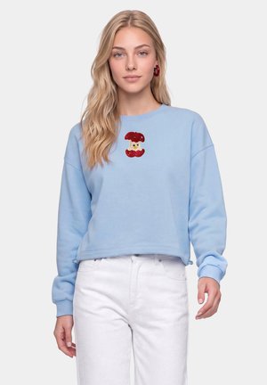 Ung kvinde med langt blondt hår iført en lyseblå sweatshirt med et rødt æblekern-design og hvide bukser mod en ensfarvet baggrund.