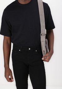 Hombre vestido con una camiseta negra y jeans, sosteniendo la correa de una bolsa de hombro de tela gris contra un fondo blanco.