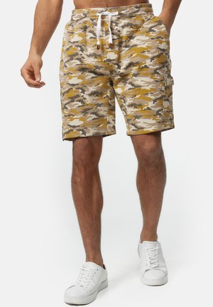 Mann trägt beige-braun-weiße Camouflage-Cargoshorts mit weißem Kordelzug und weißen Turnschuhen, steht vor einem einfarbigen Hintergrund.