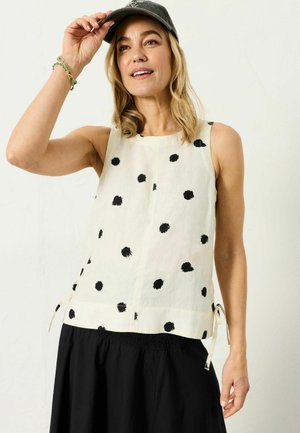 Frau trägt ein ärmelloses weißes Oberteil mit schwarzen Punkten, einen schwarzen Rock und eine dunkle Mütze, hebt die Mütze mit einer Hand an und lächelt leicht.