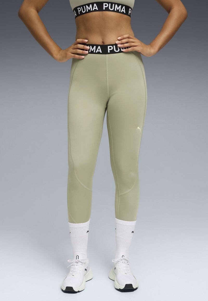 Legginsy Puma w kolorze jasnej zieleni, dopasowane do sylwetki z przeszyciami. Czarna elastyczna talia z logo "PUMA", zestawione z białymi sneakerami.
