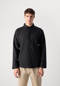 Svart langermet skjorte med knapper foran, klassisk krage og subtil logo-detalj. Glatt stoff med en tettsittende design, kombinert med beige bukser.