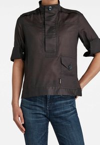 Chemise noire à manches courtes avec col montant, patte de boutonnage à l'avant et grande poche à boutons portée avec un jean bleu.