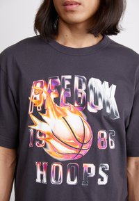Juodas medvilninis marškinėliai su grafiniu spausdiniu, kuriame pavaizduota liepsnojanti krepšinio kamuolys ir užrašas "REEBOK 1986 HOOPS" metalizuotais ir spalvotais šriftais.