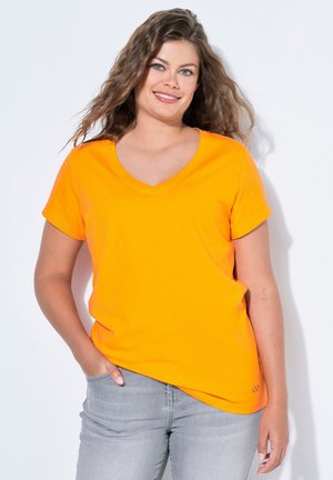 Frau mit langen, welligen Haaren, die ein leuchtend oranges V-Ausschnitt-T-Shirt und hellgraue Jeans trägt, lächelnd mit einer Hand in der Tasche vor weißem Hintergrund.