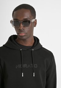 Occhiali da sole con una montatura grigia trasparente e spessa e lenti scure; indossati con una felpa nera con cappuccio caratterizzata da un logo in rilievo e dettagli argentati.