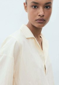 Light beige, collared shirt with a relaxed fit and subtle sheen. Features short sleeves and a half placket. Smooth texture, minimal design.