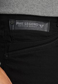 Čierne deninové džínsy s etiketou PME Legend, ktorá obsahuje šedú koženú nášivku. Obsahujú strieborný gombík a biele prešívanie pozdĺž vrecka.