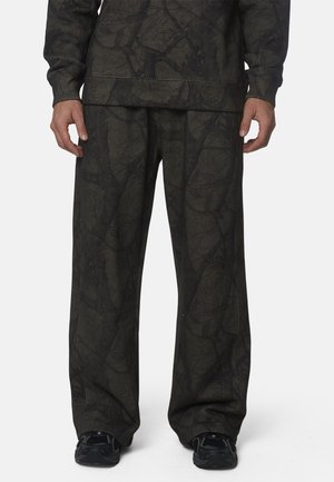 Uomo che indossa pantaloni scuri e larghi e una camicia a maniche lunghe abbinata con motivo astratto a foglia, in piedi con scarpe da ginnastica nere visibili.