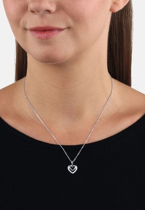 Collana con pendente a forma di cuore in argento, con un cuore nero interno, su una sottile catena, su una maglia scura, che mette in risalto il suo design a strati.