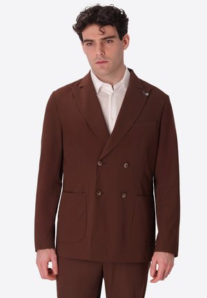 Uomo con giacca doppiopetto marrone e camicia bianca, in piedi davanti a uno sfondo chiaro uniforme.