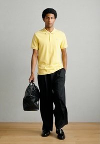 Κίτρινο πόλο πουκάμισο, άνετο μαύρο παντελόνι και μαύρα παπούτσια. Κρατάει μια υφασμένη μαύρη τσάντα χειρός. Απλός σχεδιασμός, κοντά μανίκια και άνετη εφαρμογή.